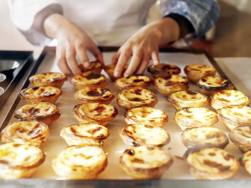 Ein Bäcker arrangiert frisch gebackene, goldbraune Pastéis de Nata auf einem Backblech in einer Konditorei in Lissabon.